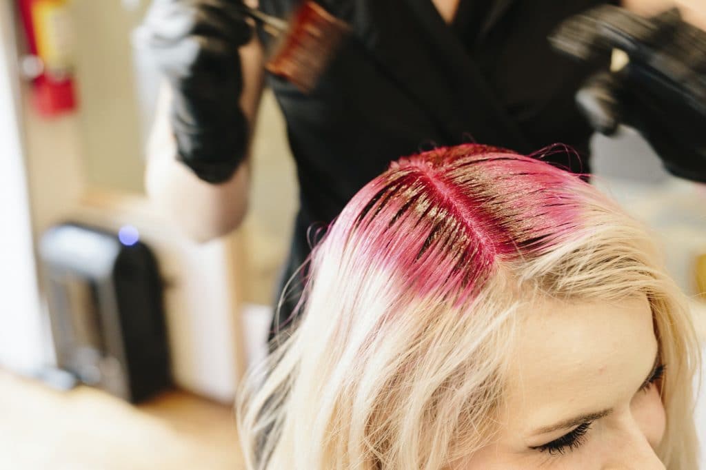 A hair colourist in gloves applying red hair dye to a client's blonde hair with a brush.